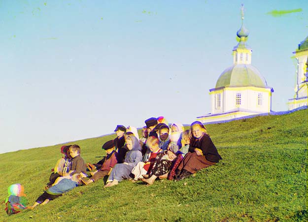 Russian children sit on the side of a hill near a church and bell tower near White Lake, in Russia, 1909.